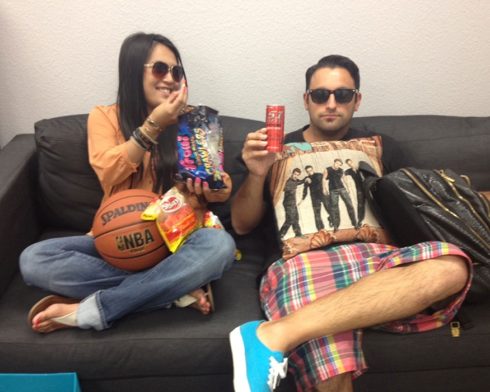 Joann Mercado Hicks and Joshua Krause relax on a couch with a basketball, gummy candy, and a boy-band pillow, smiling and enjoying a break.