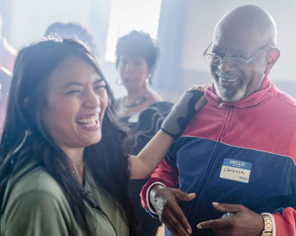 Joann Mercado Hicks laughing with a smiling gentleman named Clarence, who wears a red-and-blue tracksuit and a name tag.