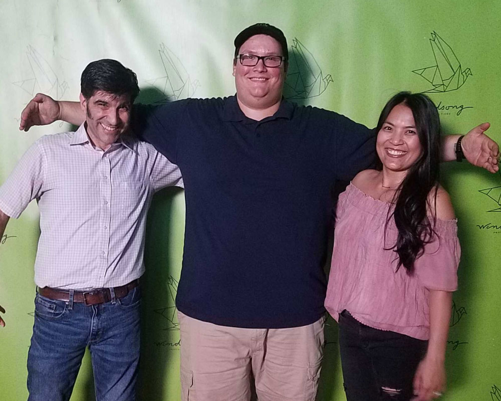 Joann Mercado Hicks standing with two colleagues in front of a green Windsong Productions backdrop.