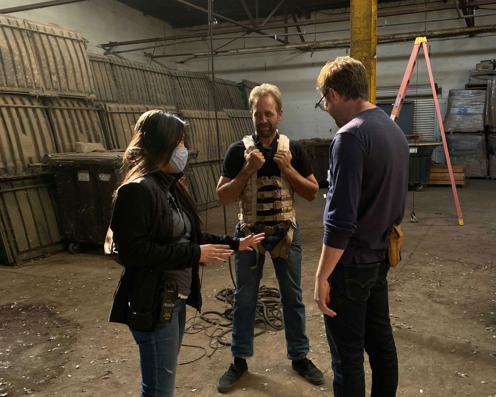 Joann Mercado Hicks, wearing a protective face mask, confers with two team members in a warehouse set about a tactical vest for a scene.