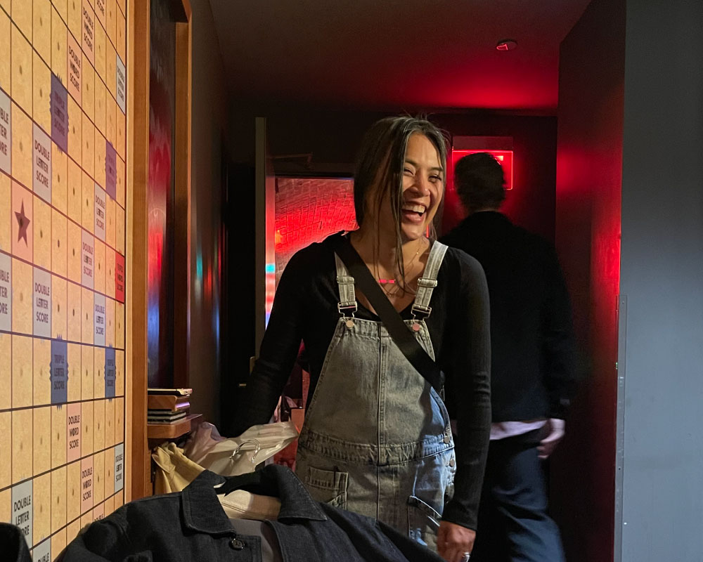 Joann Mercado Hicks in denim overalls, laughing behind the scenes near a life-size Scrabble board at a Windsong Productions shoot.