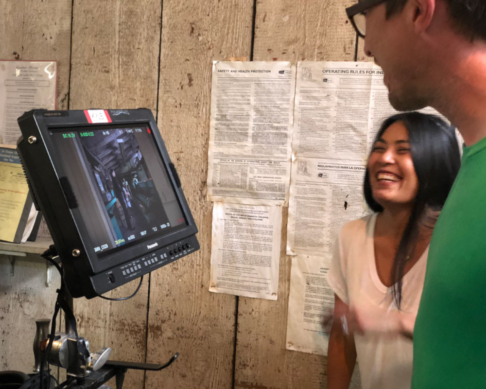 Joann Mercado Hicks grins while reviewing a camera monitor alongside a colleague in a wood-paneled workshop, discussing footage for Windsong Productions.