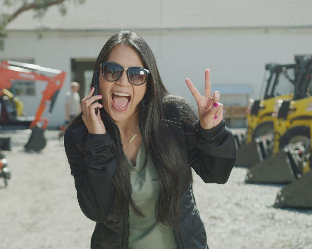 Joann Mercado Hicks, wearing sunglasses and on a phone call, flashes a vibrant peace sign in an outdoor equipment yard.