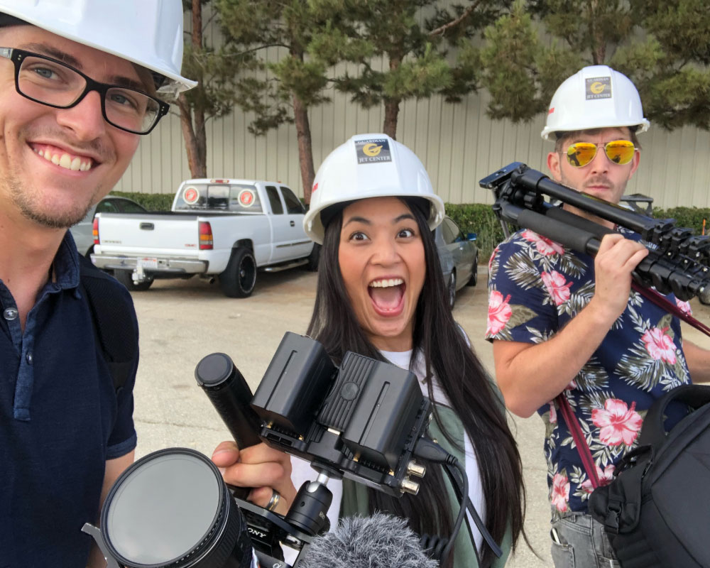 Joann Mercado Hicks in a hard hat, beaming with excitement as she holds film equipment with two colleagues on location for a Windsong Productions shoot.