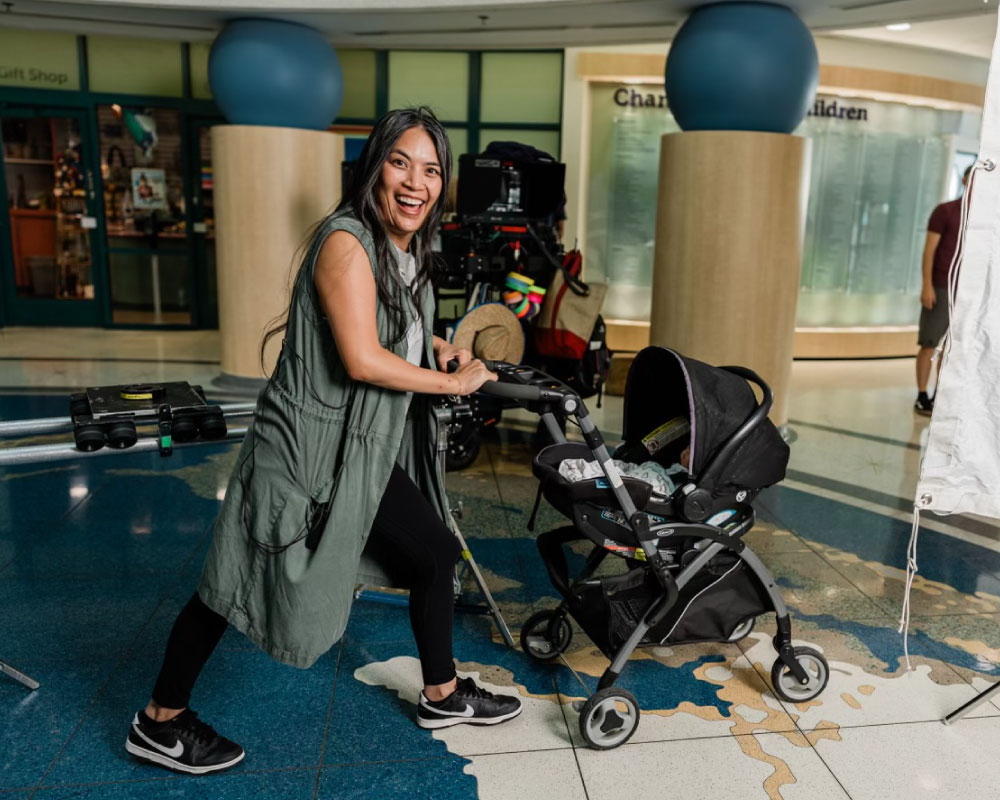 Joann Mercado Hicks laughing as she pushes a baby stroller on a busy film set inside a children’s hospital, showcasing her energetic, can-do spirit.