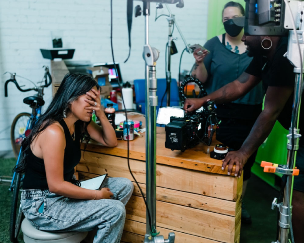 Joann Mercado Hicks, seated on set with her face in her hand, listening intently to teammates adjusting camera gear for a Windsong Productions shoot.