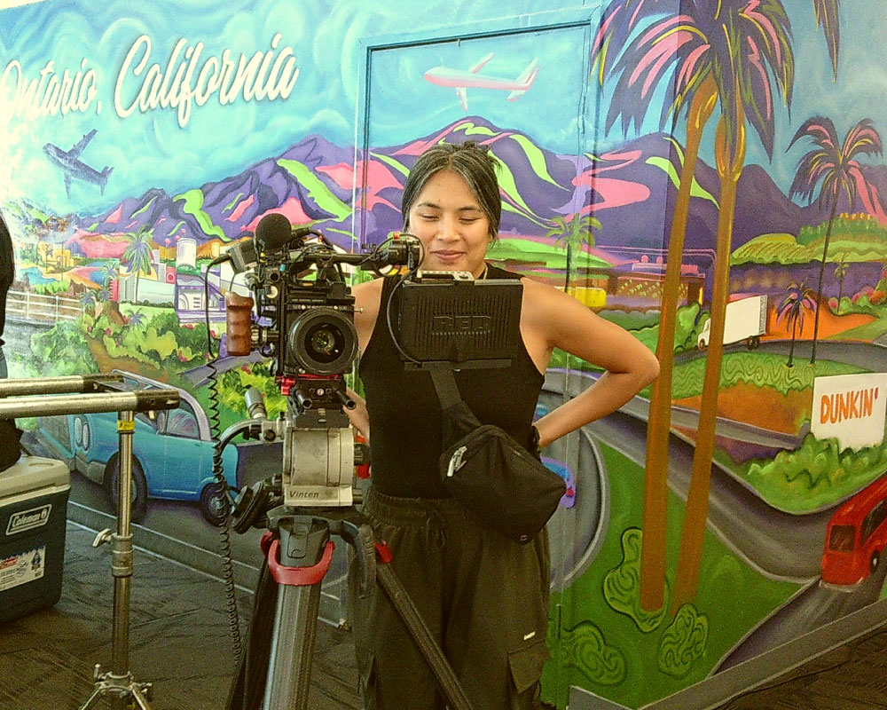 Joann Mercado Hicks standing behind a professional camera, smiling in front of a vibrant Ontario, California mural while filming on location.