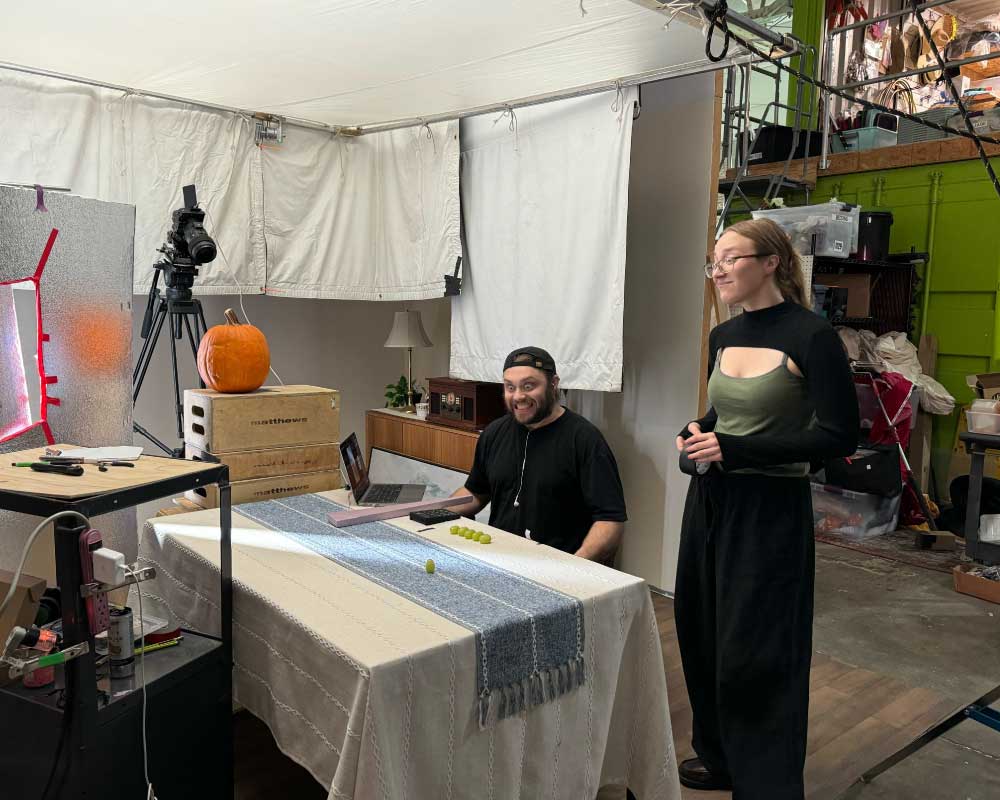 Abby Edwards, Production Assistant, and a colleague prepare for a stop-motion animation shoot. A table with a pumpkin, a laptop, and a row of green grapes sits in front of a professional camera setup, while white fabric panels diffuse light overhead.