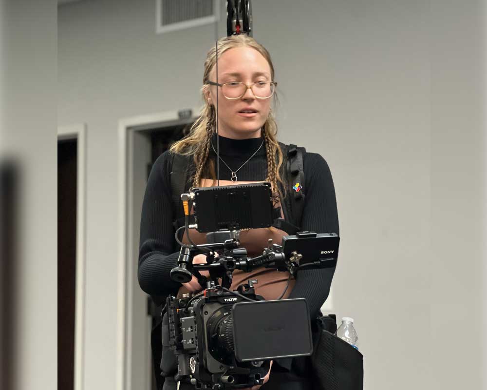 Abby Edwards, Production Assistant, operates a professional Sony camera rig with a stabilizing vest in an indoor filming environment, focused on capturing the perfect shot.