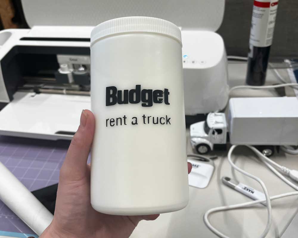 A close-up of Abby Edwards, Production Assistant, holding a white cylindrical container with a custom-made "Budget Rent a Truck" label, with a workspace full of crafting tools and materials in the background.