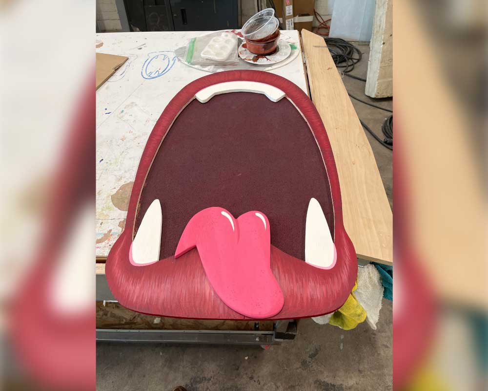 A handcrafted oversized mouth prop featuring a red tongue, white teeth, and a dark maroon background sits on a worktable, surrounded by paint supplies and tools in a production workshop.